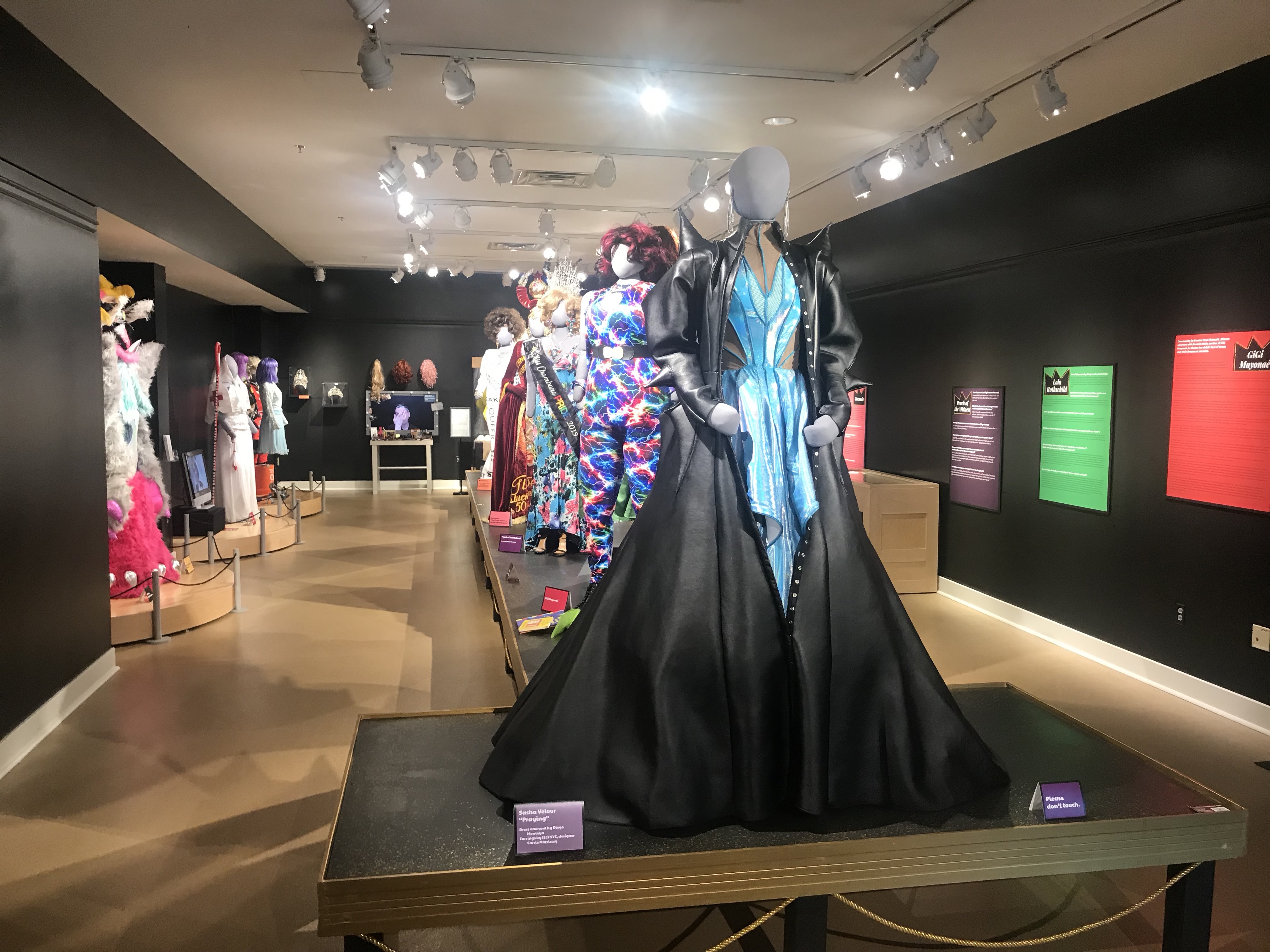 Main exhibit room for In Her Closet featuring drag costumes on grey mannequins. Text descriptions printed on multiple colored posters along black walls.
