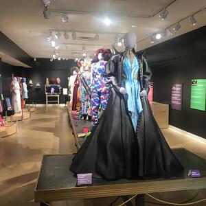 Main exhibit room for In Her Closet featuring drag costumes on grey mannequins. Text descriptions printed on multiple colored posters along black walls.